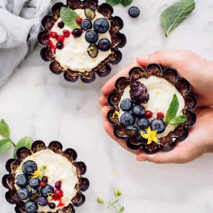 three mini lemon tarts on a white surface with fresh seasonal berries and mint
