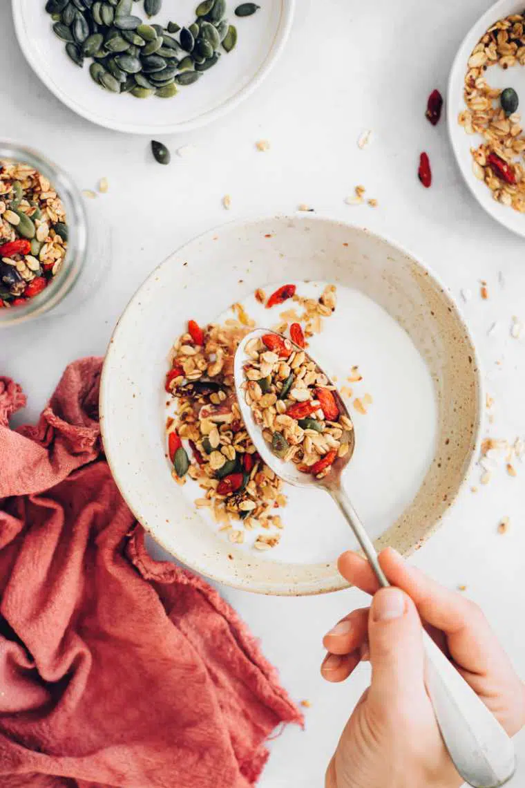 white bowl with plant-based milk and homemade vegan granola that's being sprinkled into the bowl with a spoon that's held by a hand