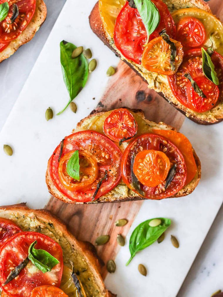 close up of three slices of baked toast with pumpkin pesto and slices of tomatoes as well as fresh basil