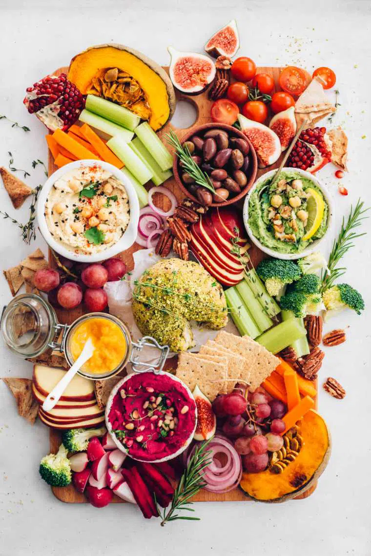 colorfully loaded board with crackers, fall produce, 3 types of hummus and nut cheese