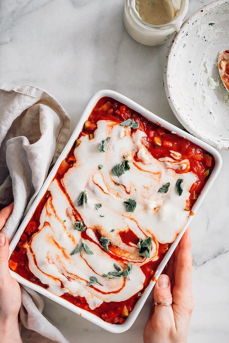 white baking dish held by two hands with tomato vegetable sauce and white dairy free cheese sauce