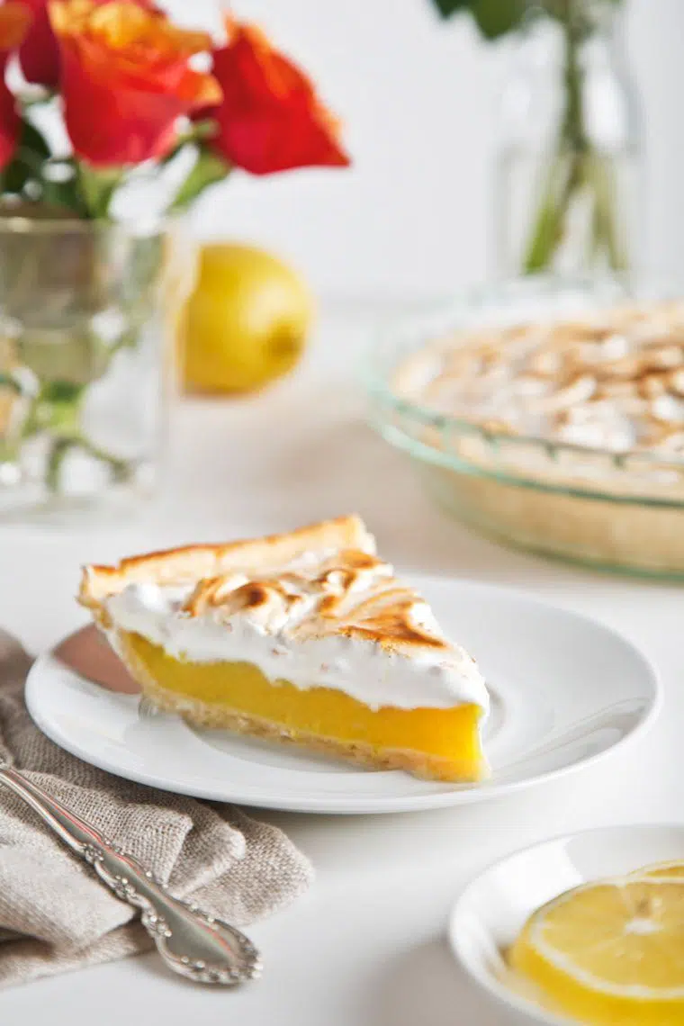 slice of vegen lemon meringue pie on a plate standing on a table with the whole pie and flowers