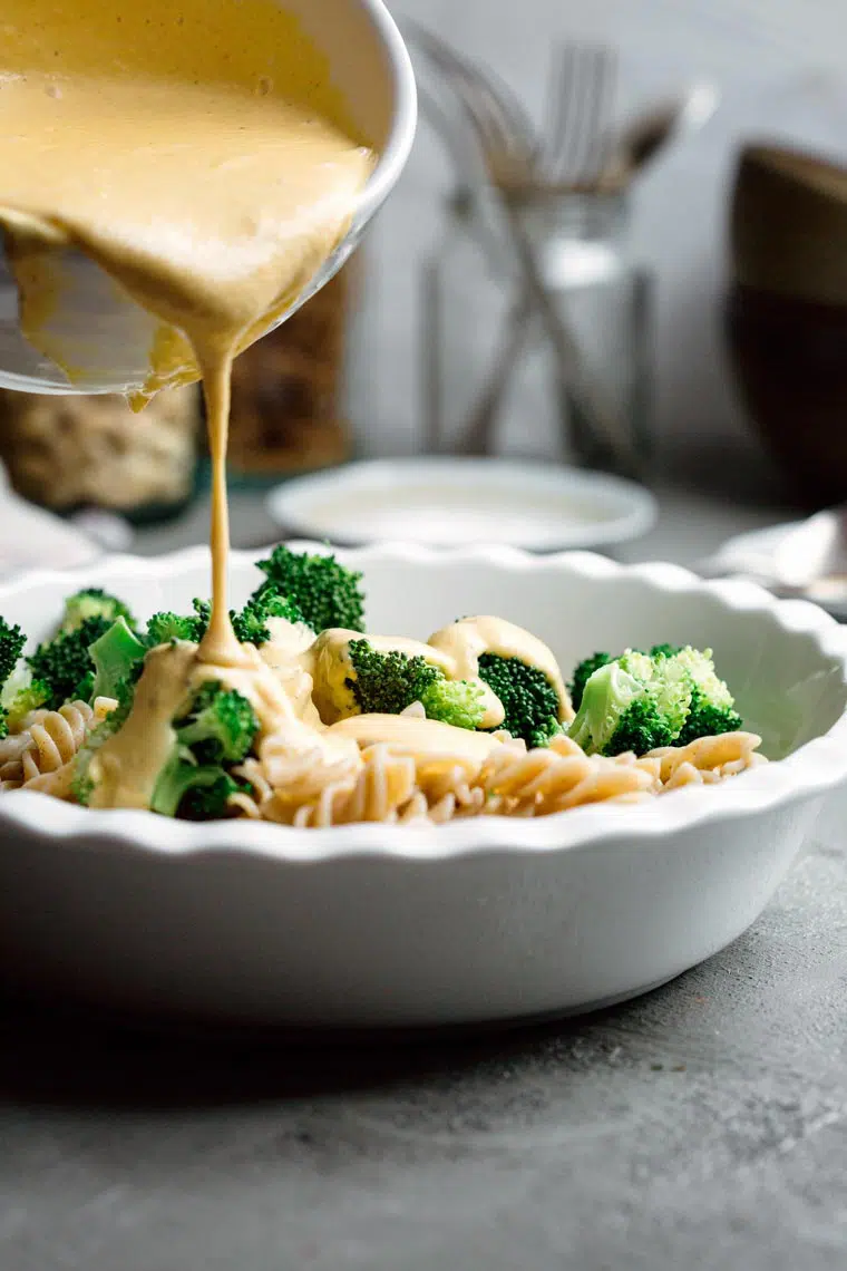 pasta and broccoli in a white bowl over which plant-based homemade cheese sauce is poured