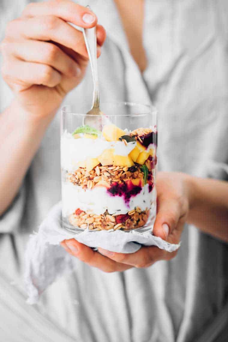 woman in white shirt holding a glass jar of homemade vegan parfait layered with vegan yogurt, granola and berries