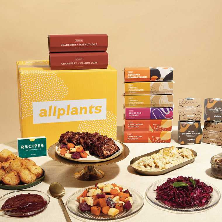 table with lots of different vegan food like cooked vegetables, potatoes, sauce and more next to food boxes by the brand allplants