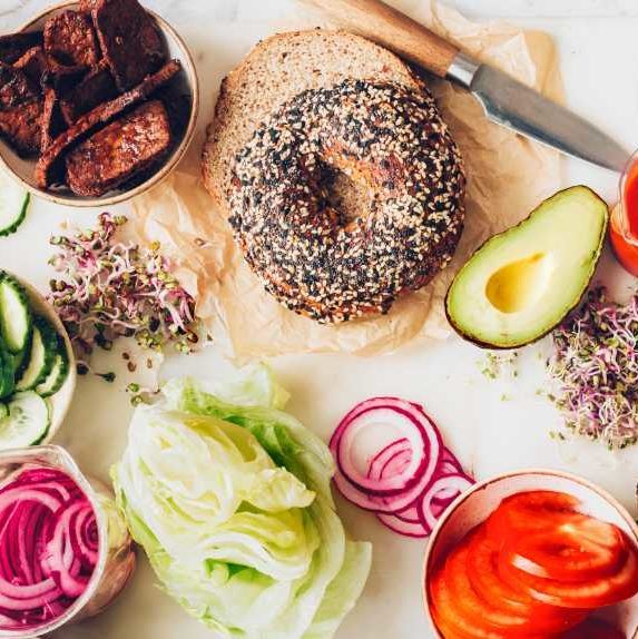 white cutting board with bagels, avocado, lettuce, red onion, tempeh and vegan cream cheese