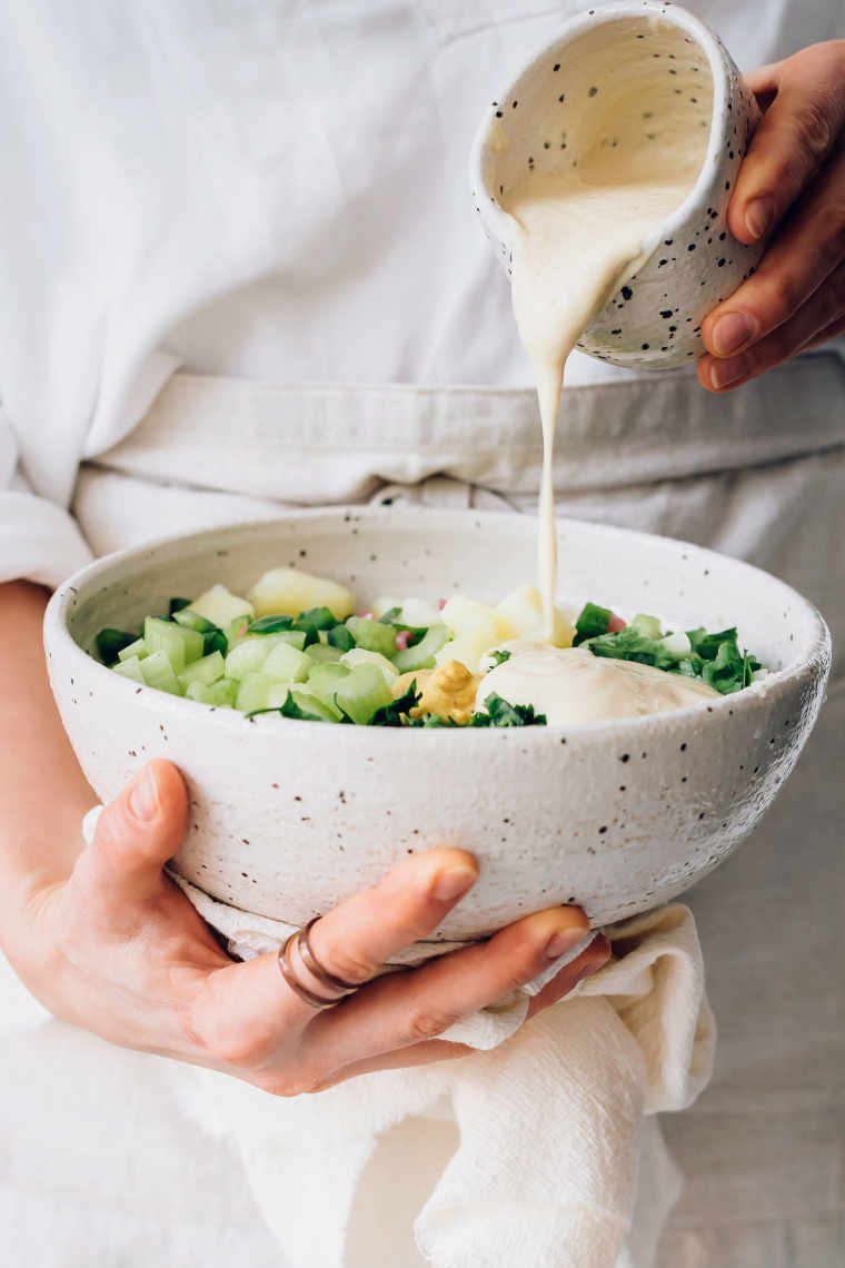 woman in linen apron holding bowl of potato salad in one hand and pouring cashew mayo over it with the other