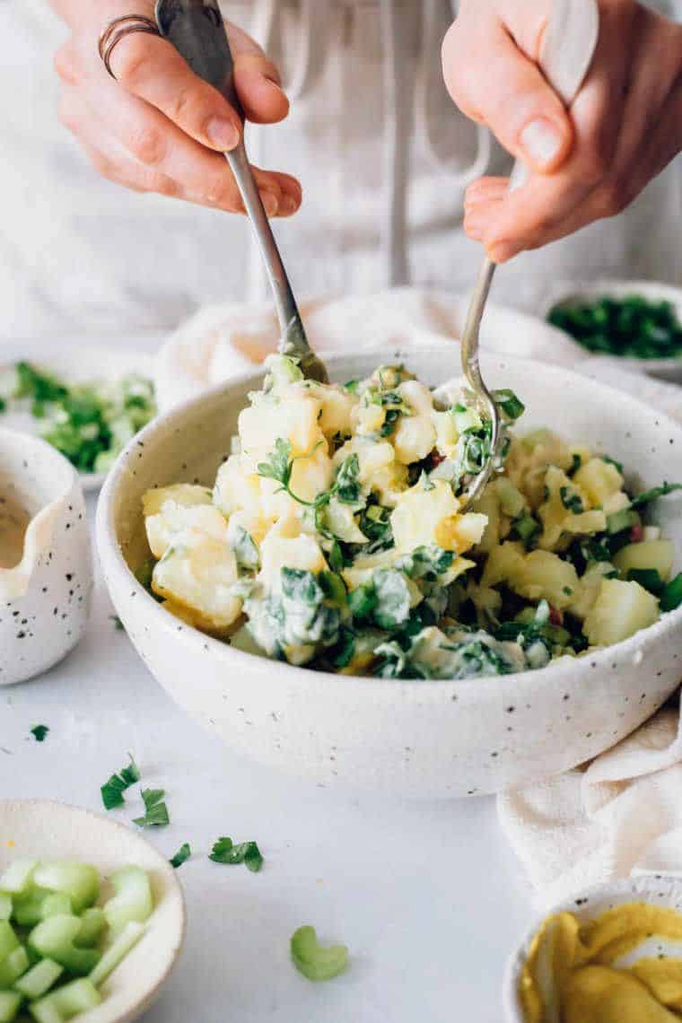 woman standing next to a bowl of potato salad and mixing it with two spoons