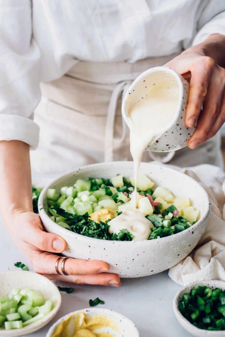 woman in white apron standing next to a bowl of potato salad and pouring cashew mayo over it