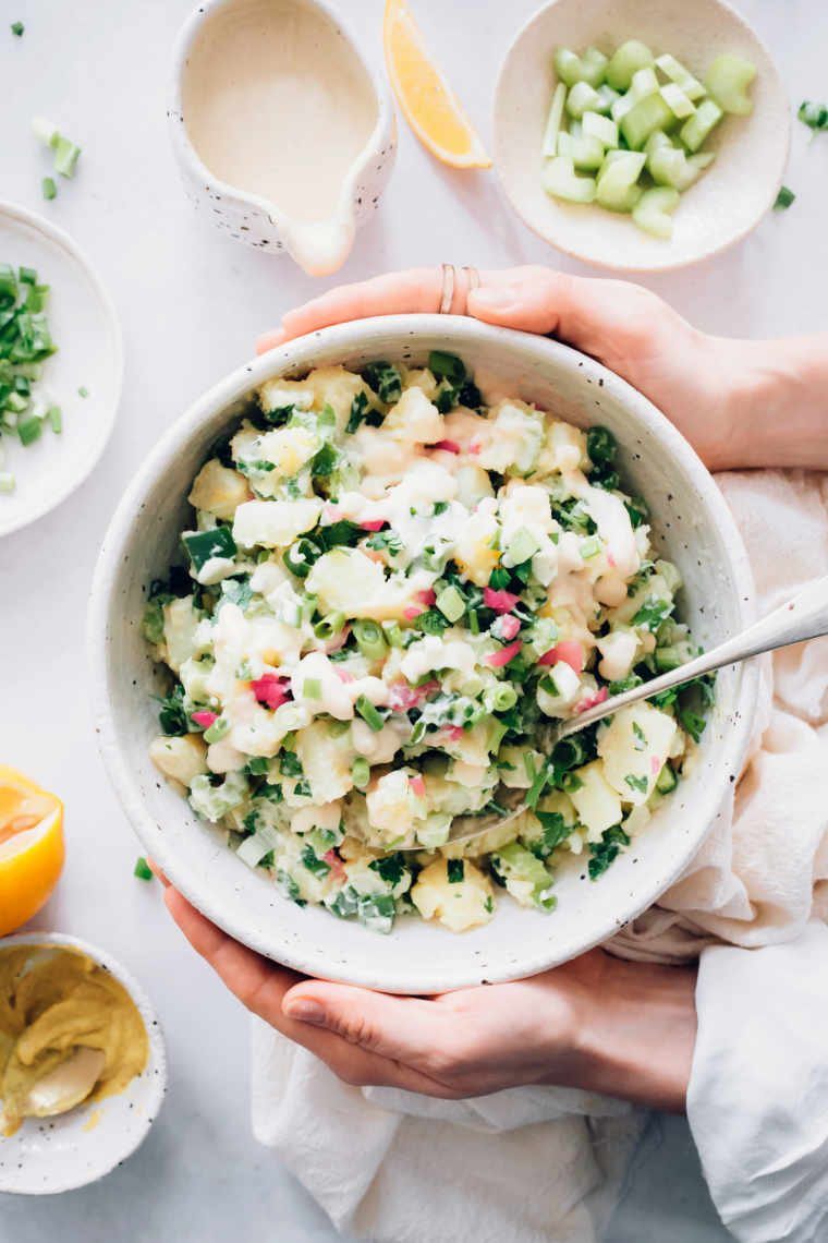 woman holding a creamy vegan potato salad over a white table with ingredients for the salad