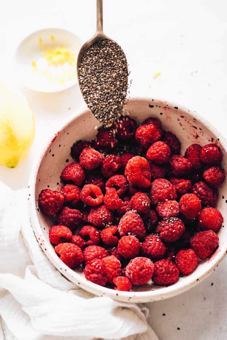 large white bowl with raspberries over which chia seeds are being sprinkled with a spoon