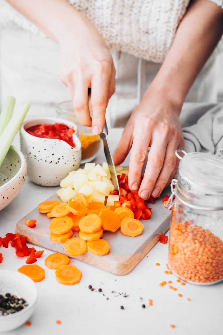 woman in white comfy sweater chopping potatoes, carrots and red bell peppers on a white board