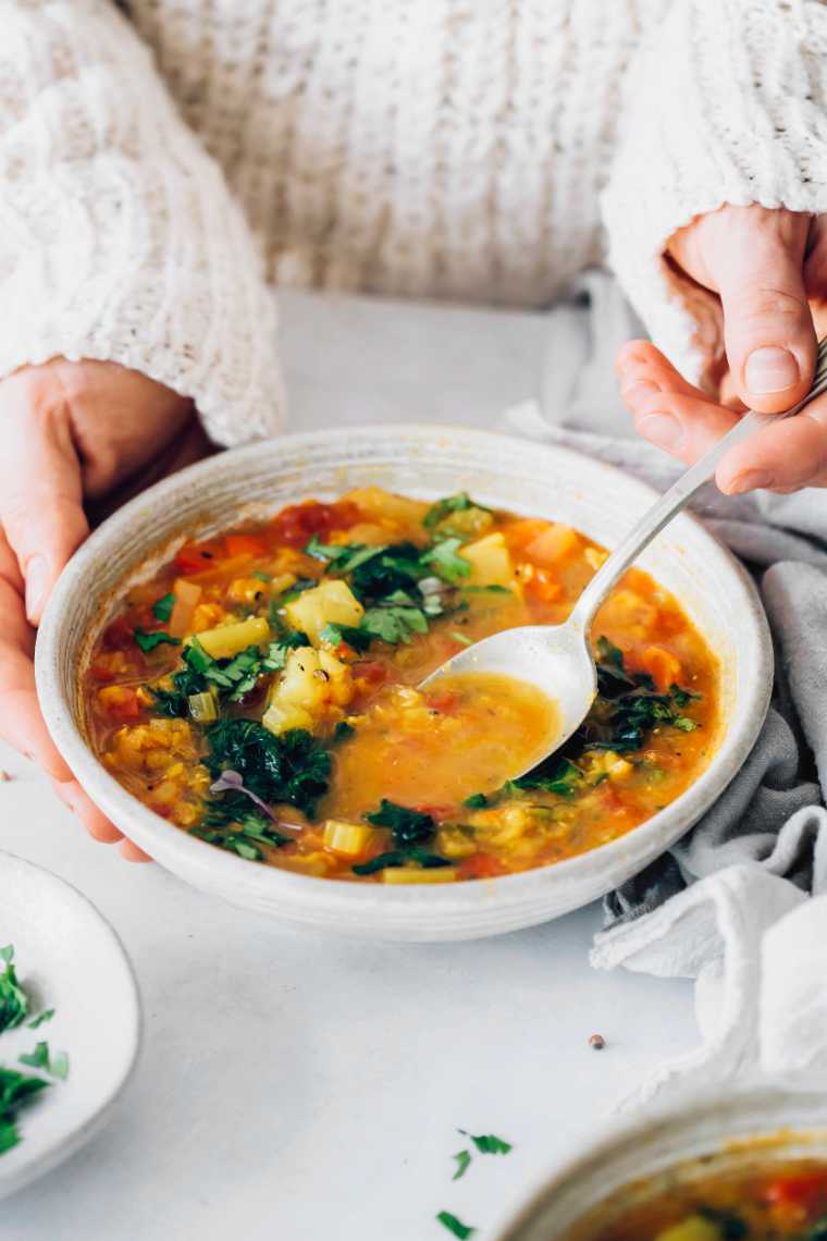 woman in cozy white sweater eating a bowl of orange anti-inflammatory soup with veggies using a spoon