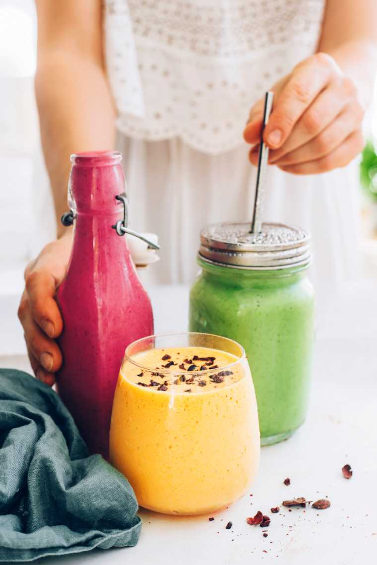 woman in white clothes standing behind three colorful vegan smoothies