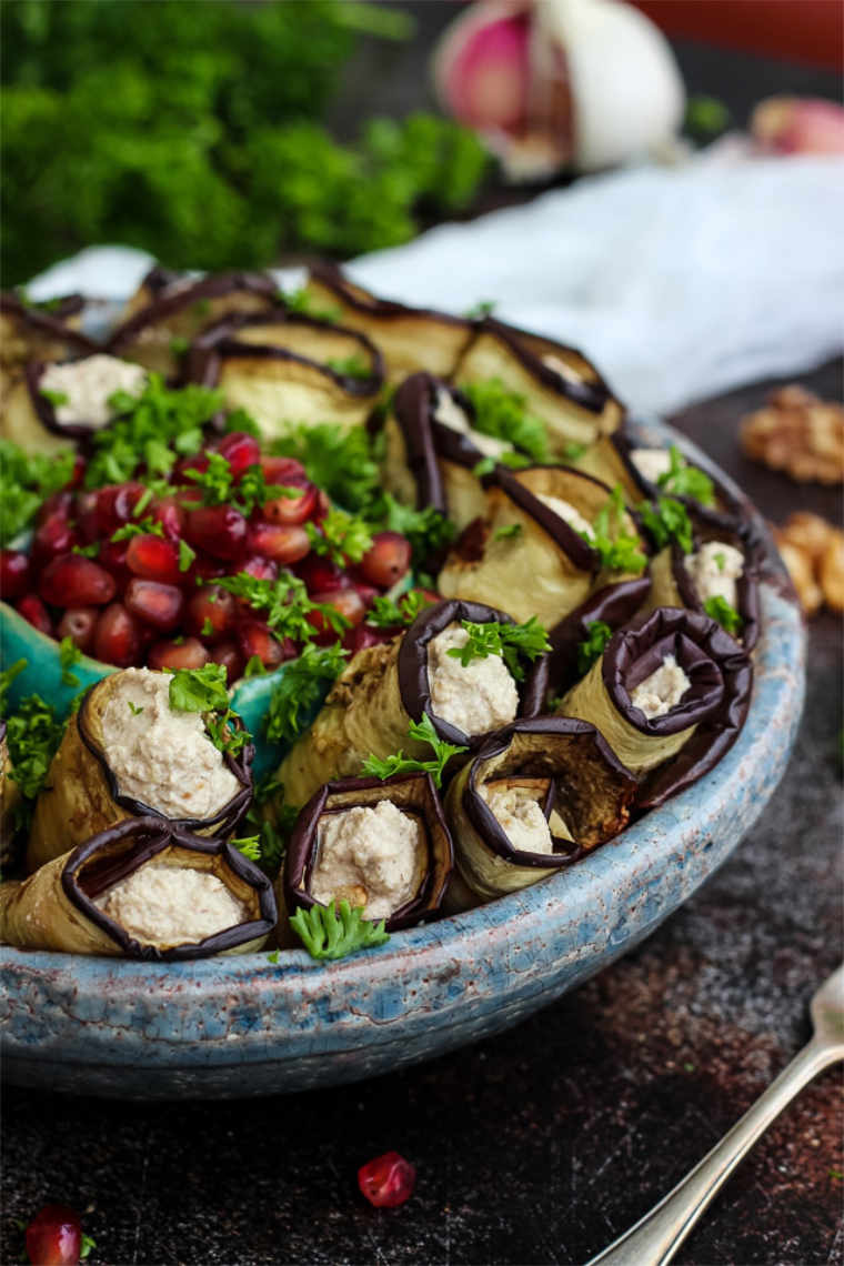 blue bowl on a table filled with vegan eggplant rolls and fresh herbs, topped with pomegranate seeds