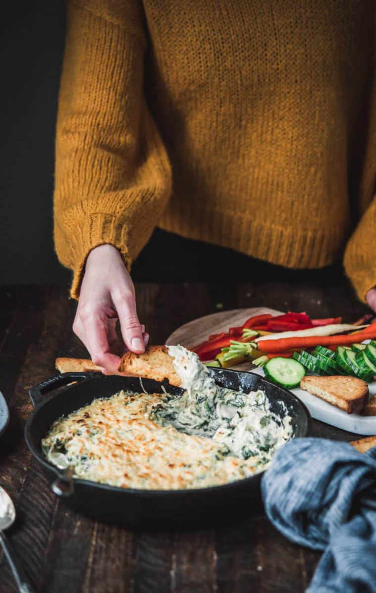 woman in yellow sweater standing at a wooden table and dipping a piece of bread into a vegan cheesy spinach artichoke dip
