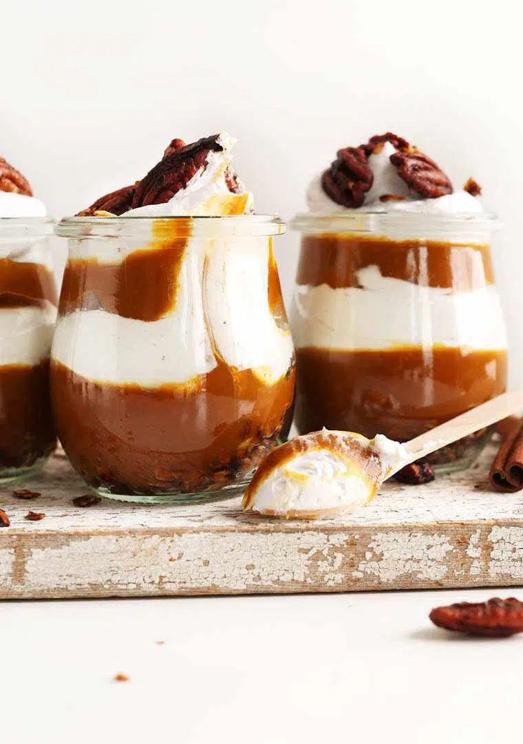 three glasses with layered pumpkin pie and vegan cream parfaits topped with pecans on a wooden surface