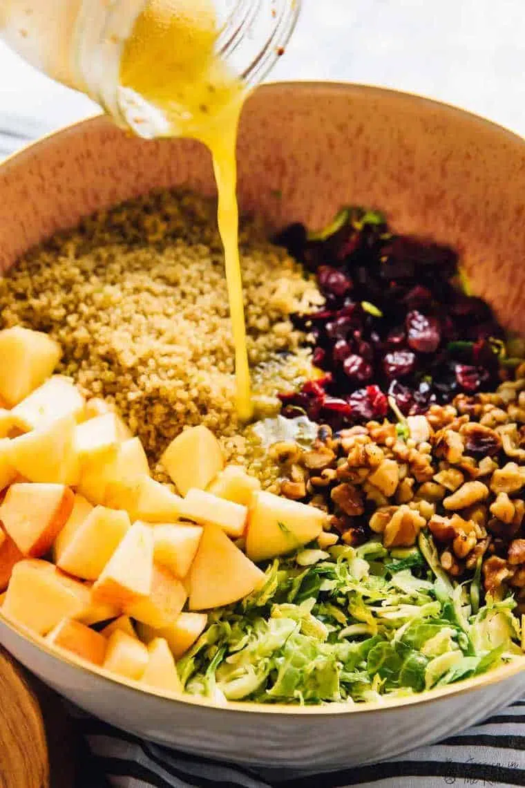 large bowl with cooked quinoa, shaved brussels sprouts, walnuts, dried berries and chopped apples being all drizzled with a yellow dressing