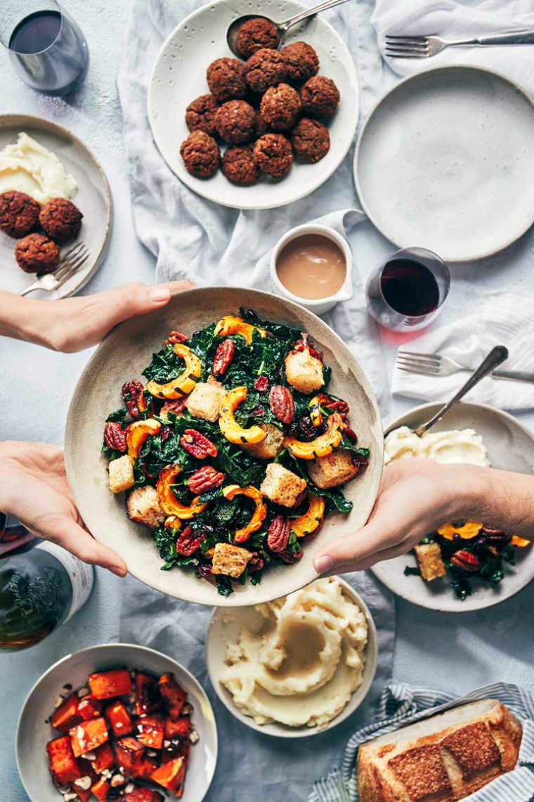 table spread with different vegan thanksgiving sides in bowls such as a kale and pumpkin salad, mashed potatoes, falafel and a loaf of bread