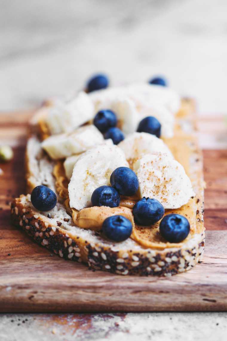 wooden chopping board with a slice of bread topped with peanut butter, banana and blueberries
