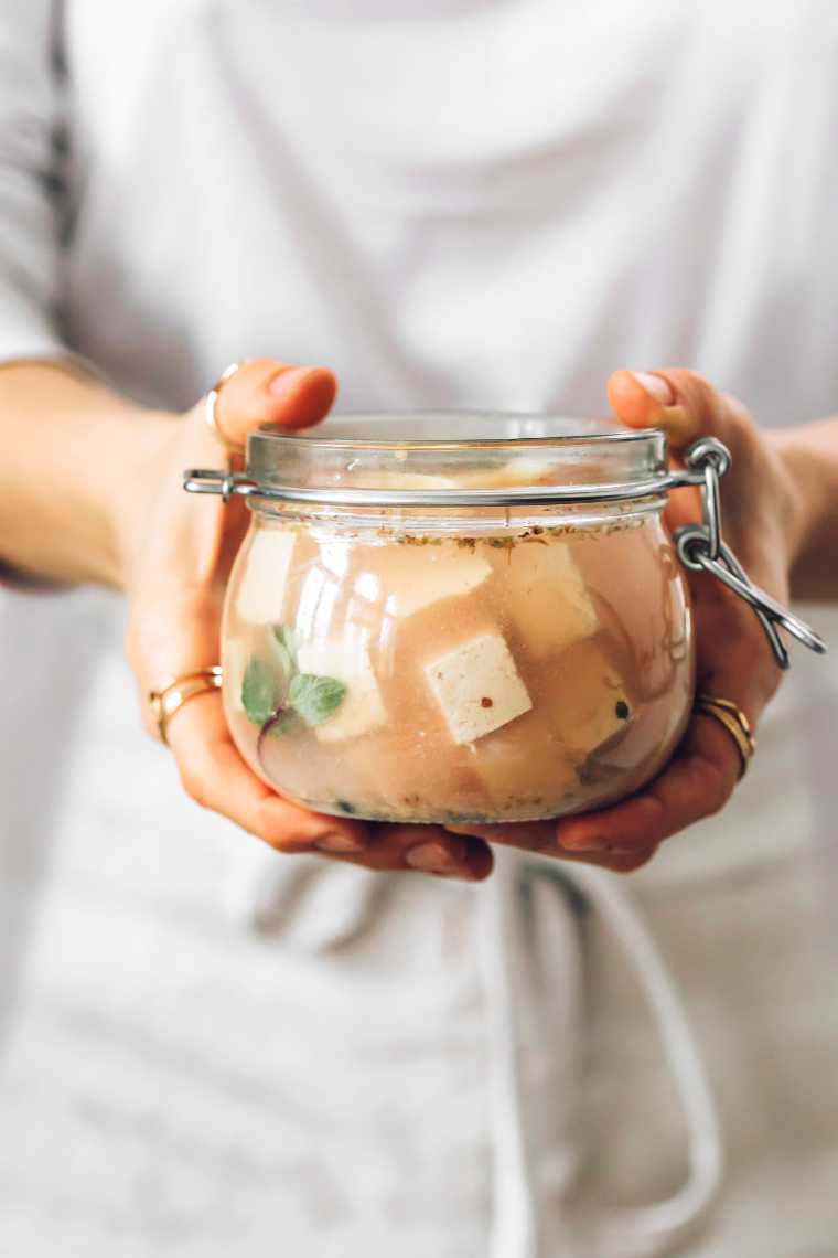 woman in white shirt and apron holding a glass jar with homemade briny vegan tofu feta and herbs