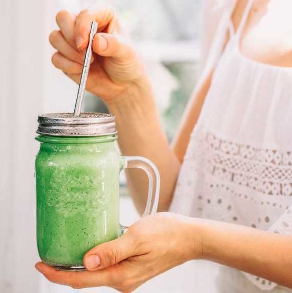 woman in white dress standing by a window and holding a glass jar with homemade green smoothie