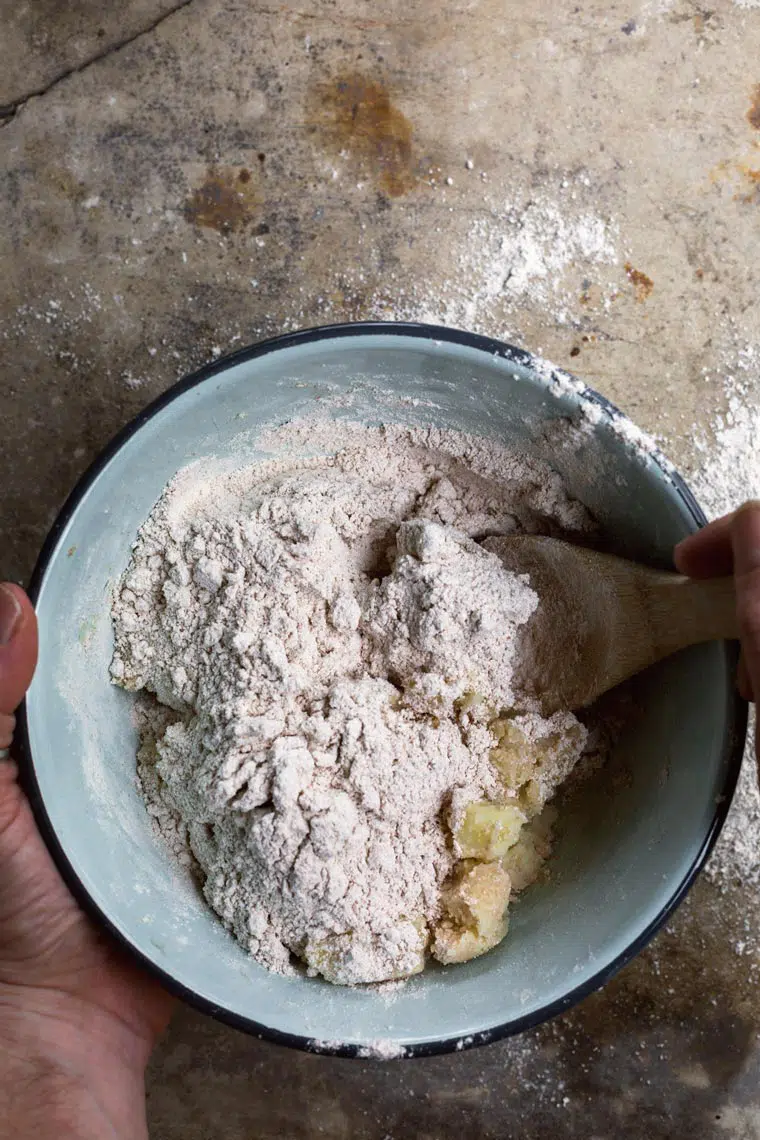 blue bowl on a floured surface in which a person mixed mashed potatoes with whole wheat flour using a wooden spoon