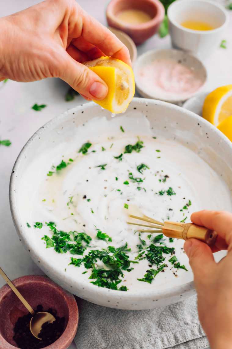 large white bowl with vegan Green yogurt dressing in which a hand sprinkles some lemon juice and whisks it