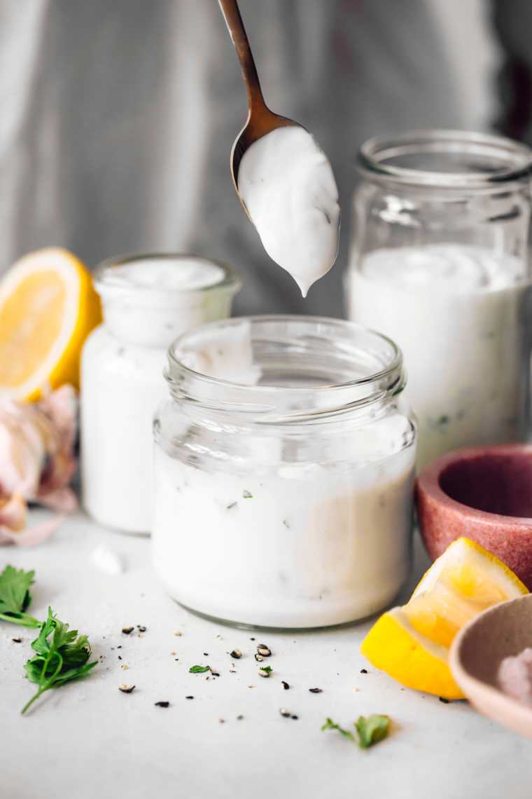 woman standing behind a table and holding a spoon with thick homemade vegan Greek yogurt dressing with garlic, tahini and parsley
