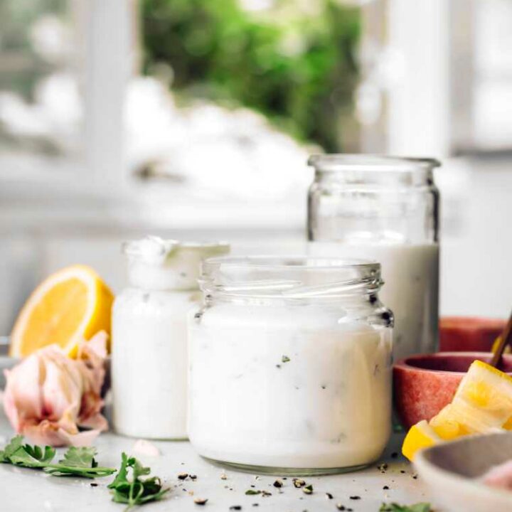 white table next to a window with three glass jars containing homemade vegan garlic yogurt sauce
