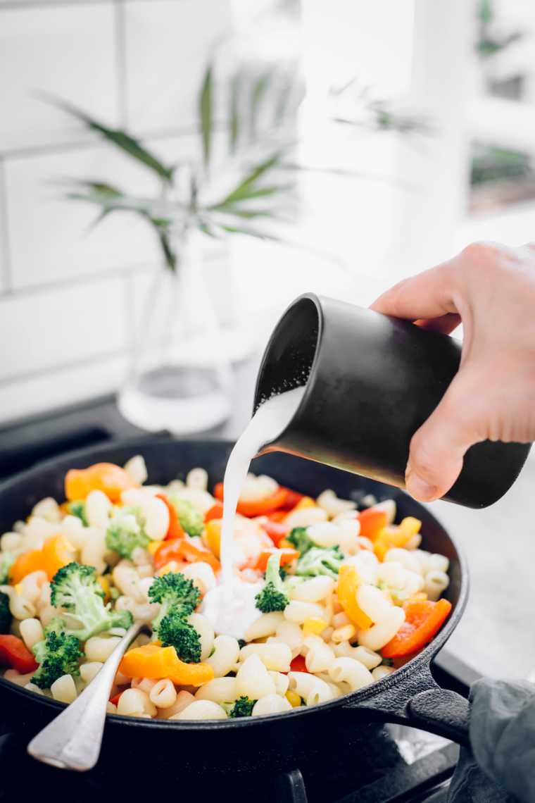 hand pouring vegan white sauce over pasta with vegetables in a pan