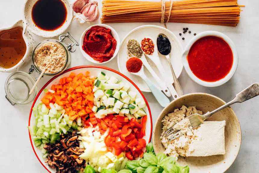 White table with whole-grain spaghetti, tofu, colorful chopped vegetables, tomato sauce and herbs