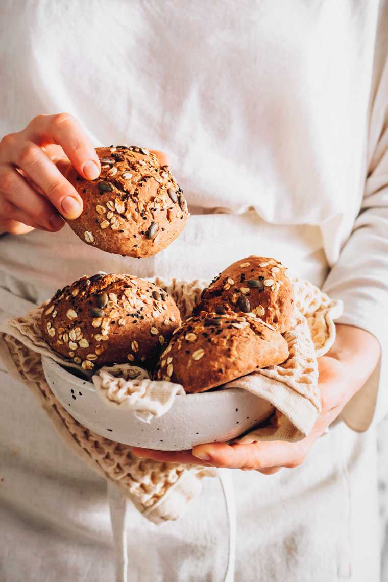 woman in white linen shirt and apron holding a bowl of freshly baked whole wheat burger buns