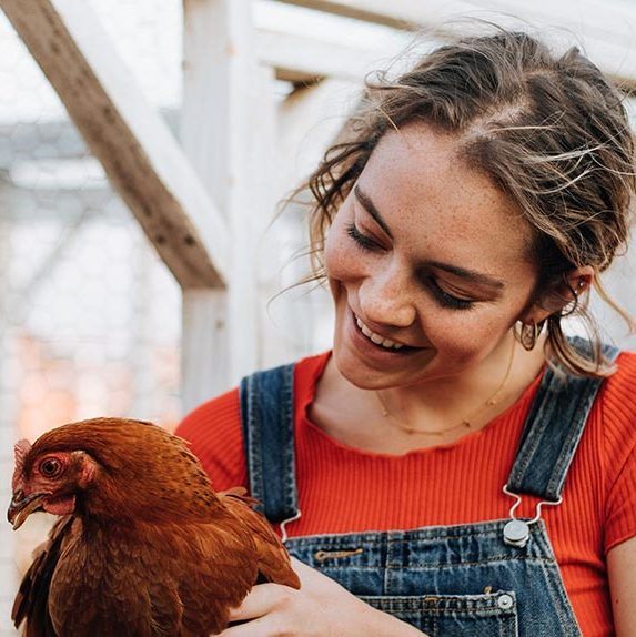 freckled woman with ponytail in red shirt and jeans holding a brown feathered chicken and smiling