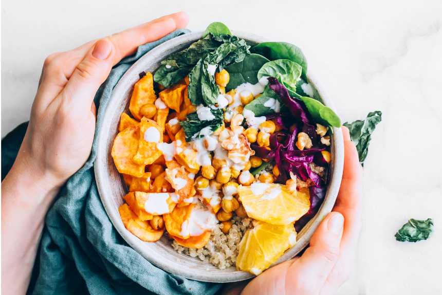 two hands holding a colorful plant-based bowl made from roasted and fresh veg, chickpeas, quinoa and dressing