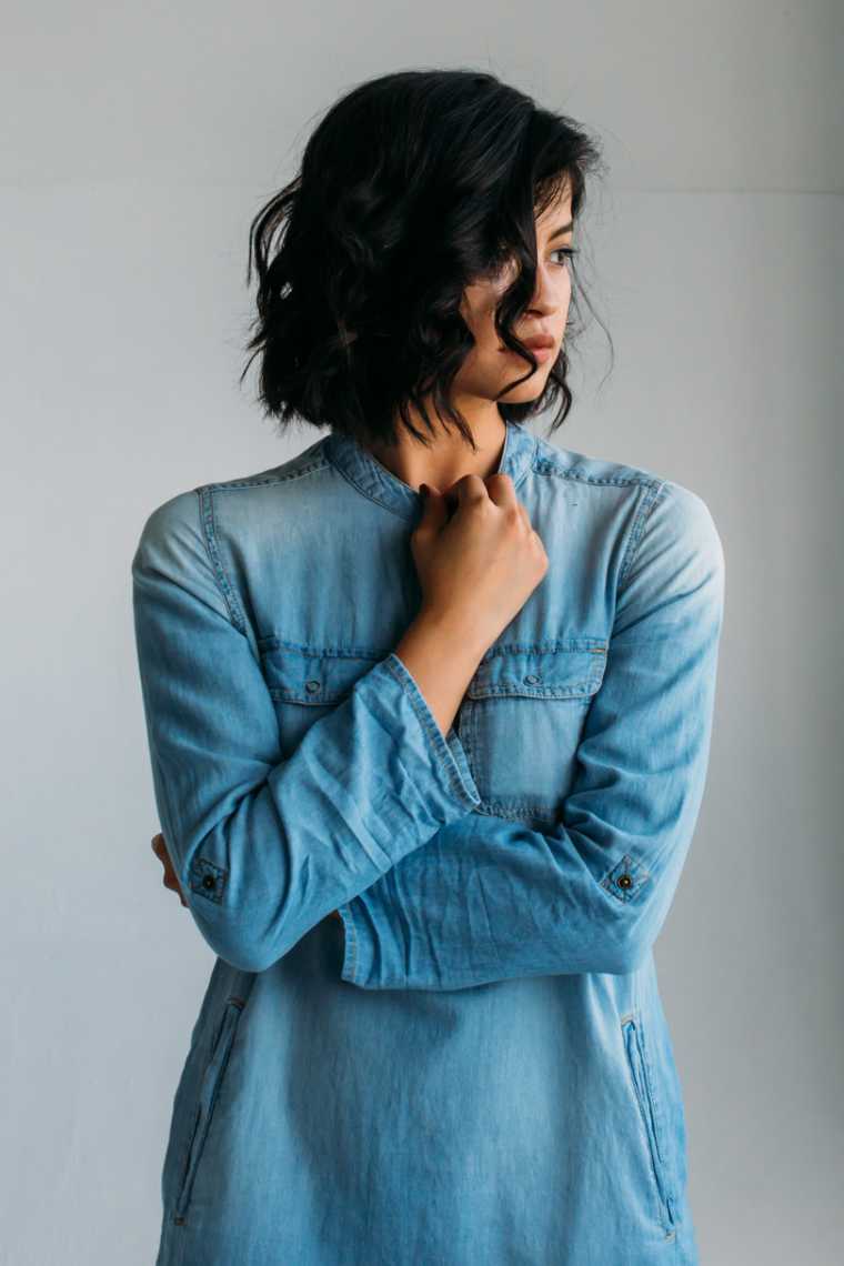 black-haired young woman wearing a jeans shirt standing in front of a grey wall and looking to her left