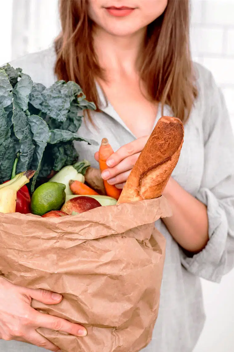 brown haired woman holding a paper bag with different vegan groceries such as bread, carrots, banana, apples and leafy greens