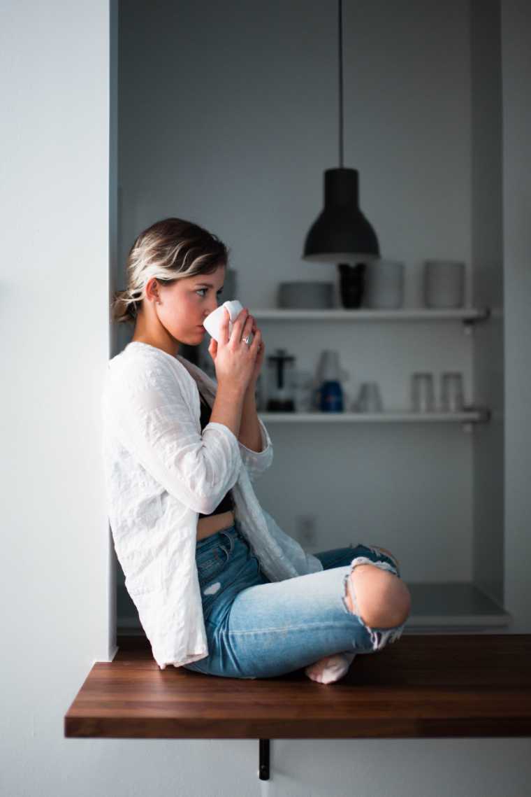 Femme blonde assise en tailleur sur un comptoir dans une cuisine minimaliste, buvant une tasse de thé