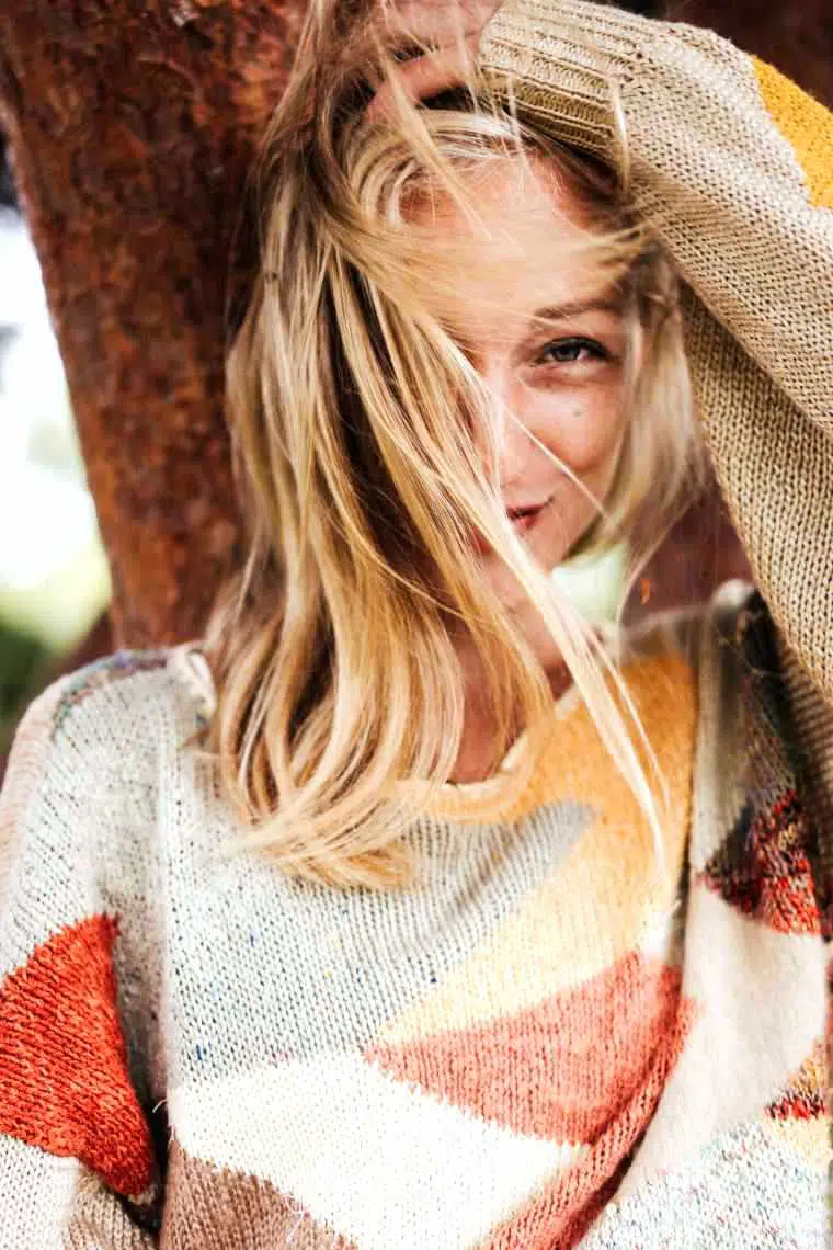 woman with blonde hair and a warm sweater standing in front of a tree and smiling
