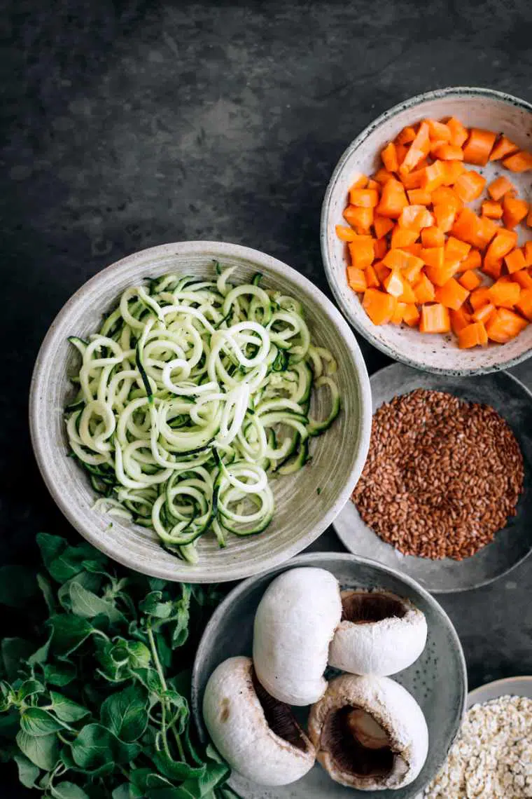 Different small bowls on a black table filled with ingredients for the zoodles with black bean meatballs recipe: zucchini, carrots, flaxseeds, mushrooms, oats and herbs