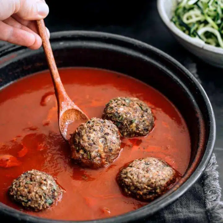 Woman using wooden spoon to place vegan meatballs into a pan filled with tomato sauce