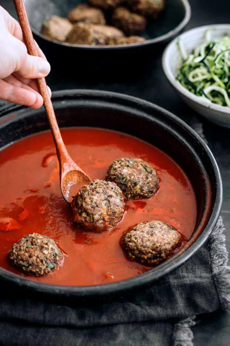 Woman using wooden spoon to place vegan meatballs into a pan filled with tomato sauce