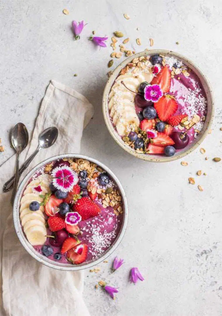 two bowls on a table filled with blueberry acai smoothie and topped with flowers, berries, banana and granola