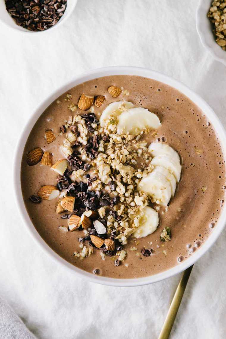white table with a bowl of brown chocolate almond butter smoothie topped with almonds, chocolate and banana