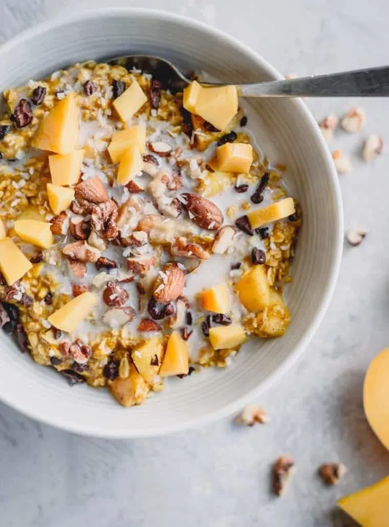 white bowl with golden oatmeal, mango, chopped nuts and vegan milk as well as a spoon