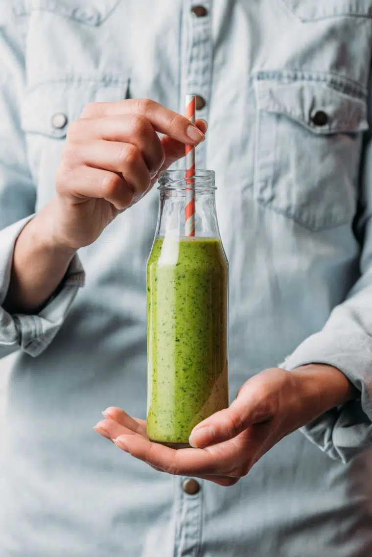 person in jeans jacket holding a tall glass with green smoothie and a straw