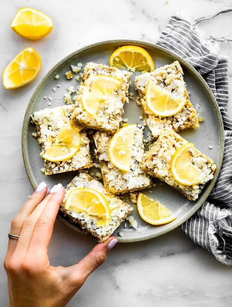 grey plate with six crumbly no-bake lemon bars topped with a slice of lemon and a hand reaching for one