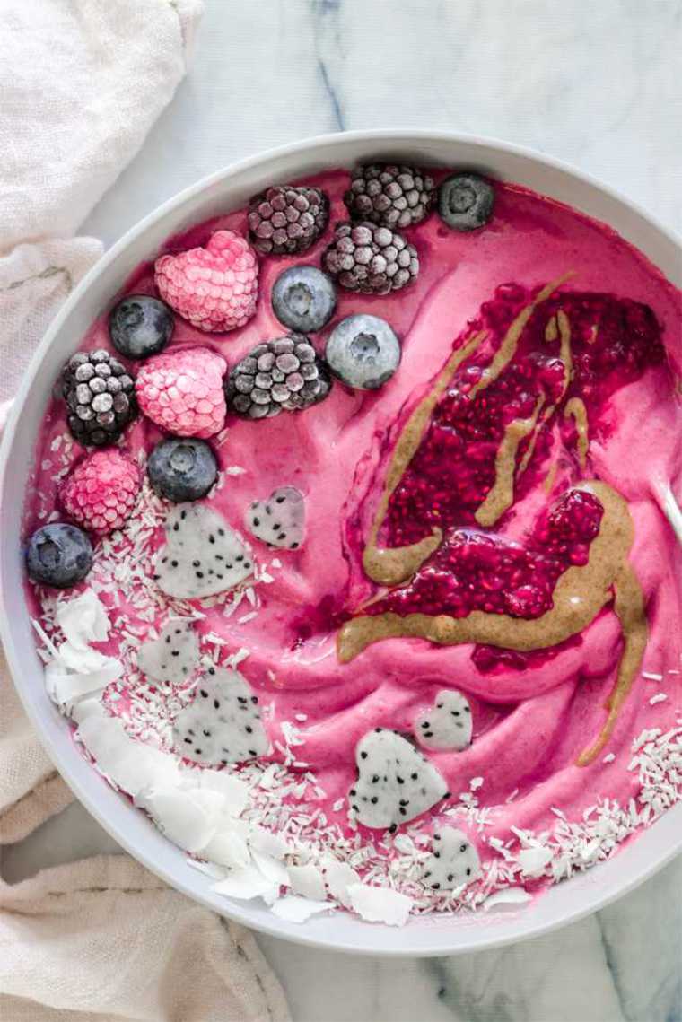 large white bowl filled with bright pink smoothie, raspberry jam, pitaya, frozen berries and coconut flakes
