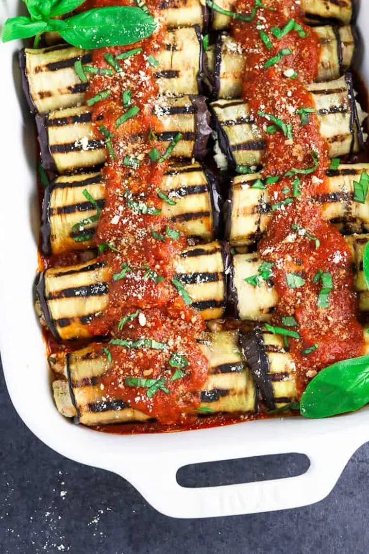 white baking pan with tomato sauce and vegan ricotta stuffed eggplant roll ups decorated with basil