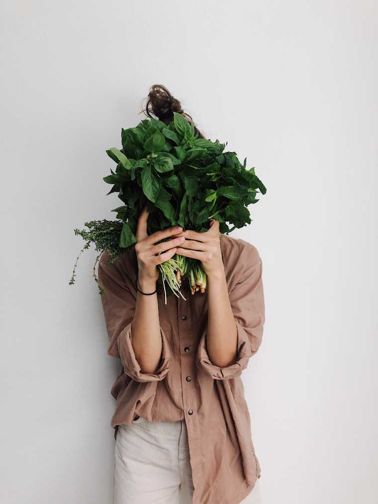woman in linen clothing standing in front of a wall and holding different leafy greens